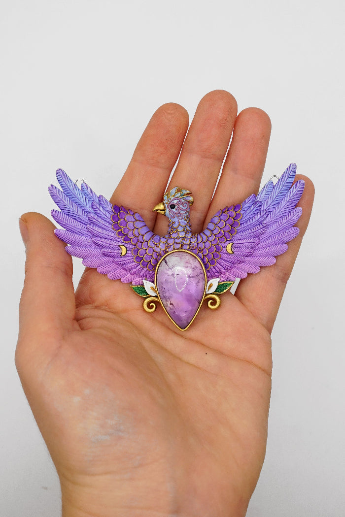 Purple gemstone pendant with wings held in a hand against a white background