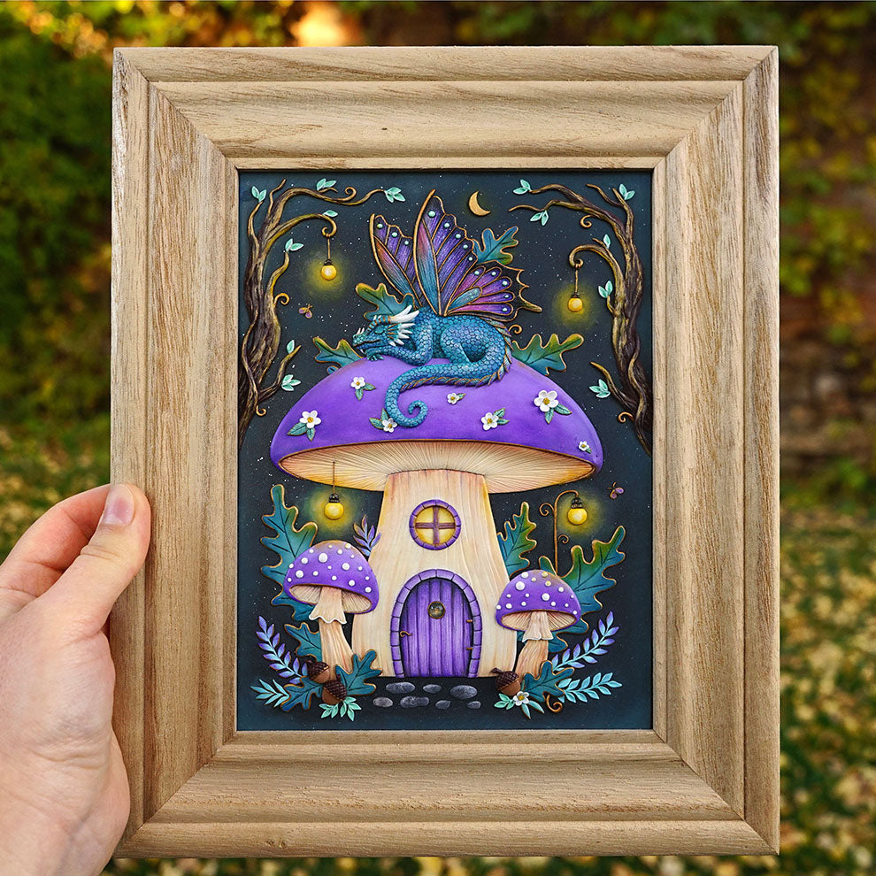 A hand holds a square wooden frame containing a colorful fantasy artwork of a blue dragon perched on a purple mushroom house, with smaller mushrooms and glowing lights around it, against an outdoor blurred green background.