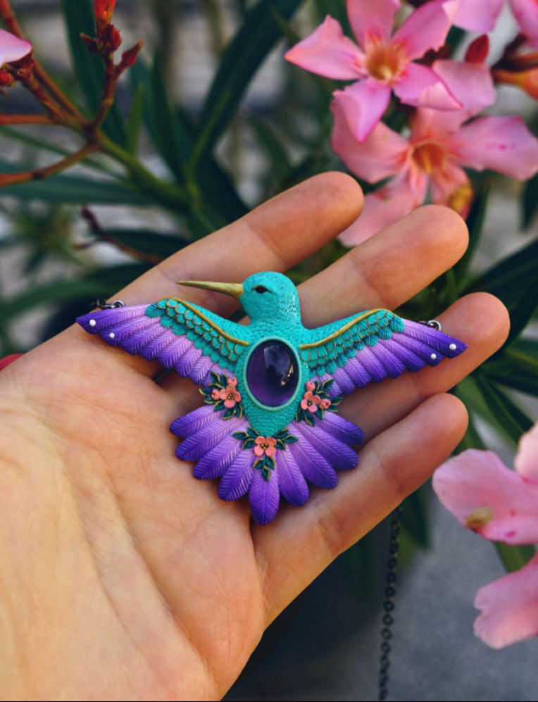 Hand holding a purple and teal bird-shaped pendant with pink flowers in the background.