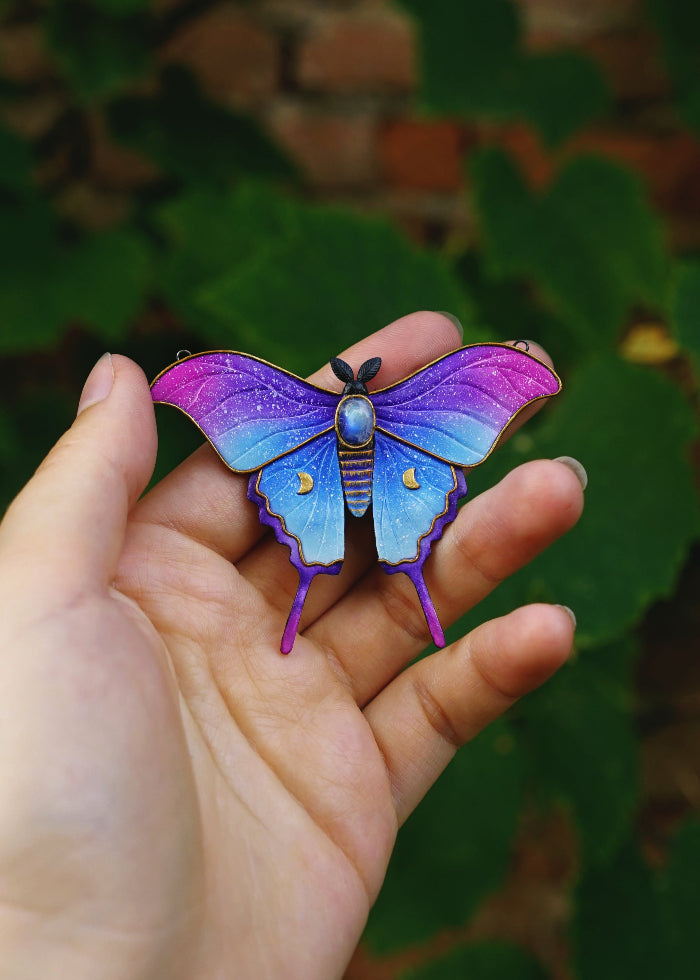 Colorful butterfly-shaped pendant held in a hand with a natural background. Made by polymer clay artist Andreea Corpodean of Wildflower Tales.