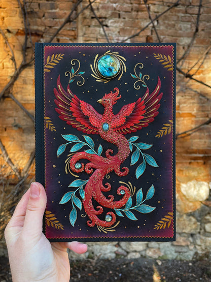 Hand holding a decorative notebook with a red phoenix design against a rustic stone wall.