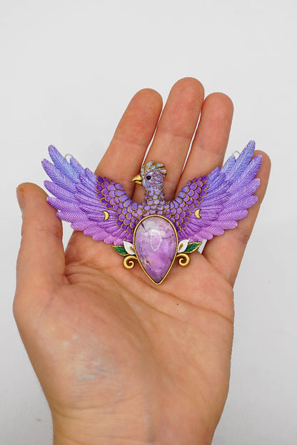Purple gemstone pendant with wings held in a hand against a white background