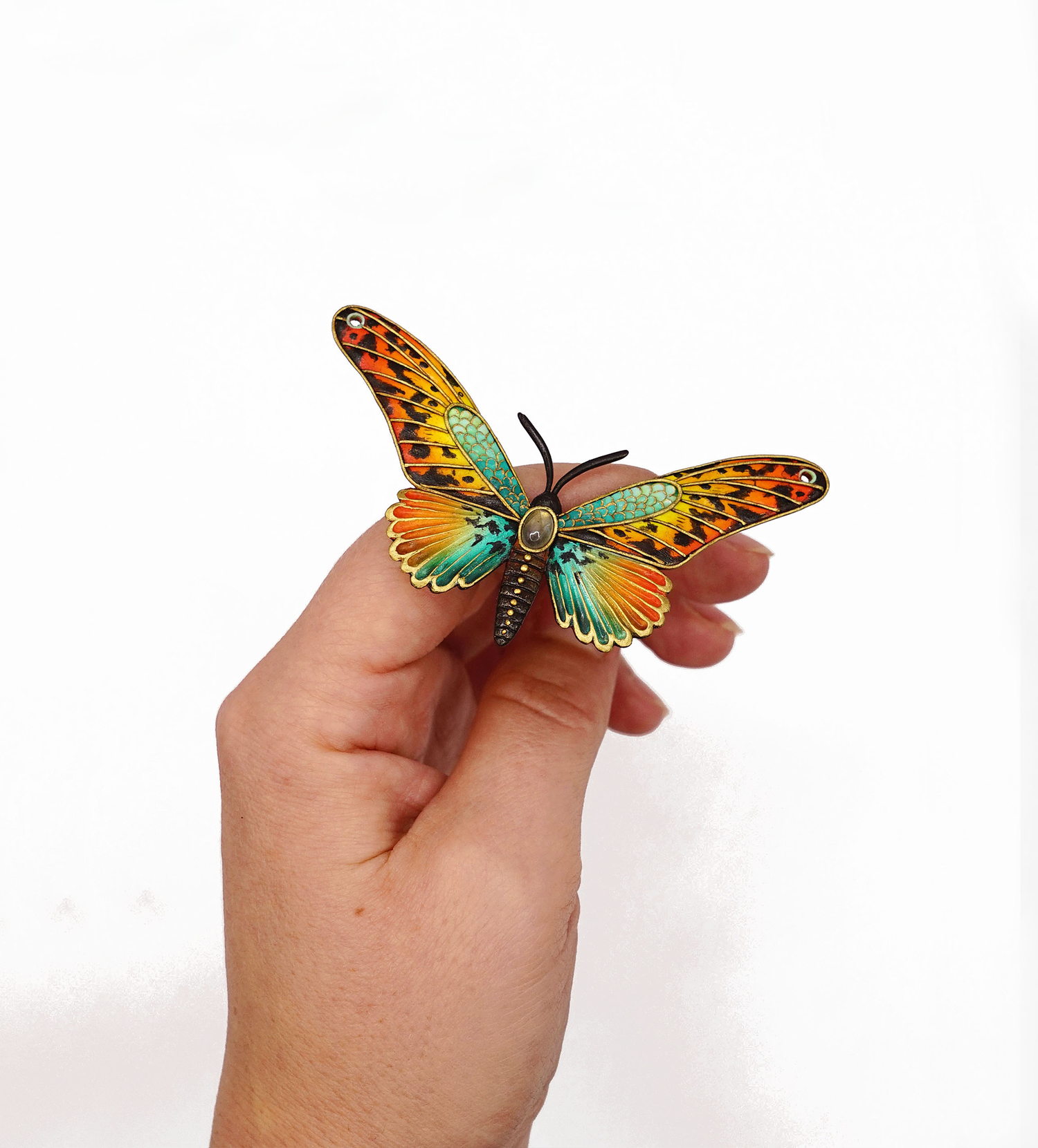 Butterfly-shaped brooch with colorful wings held in a hand on a white background.