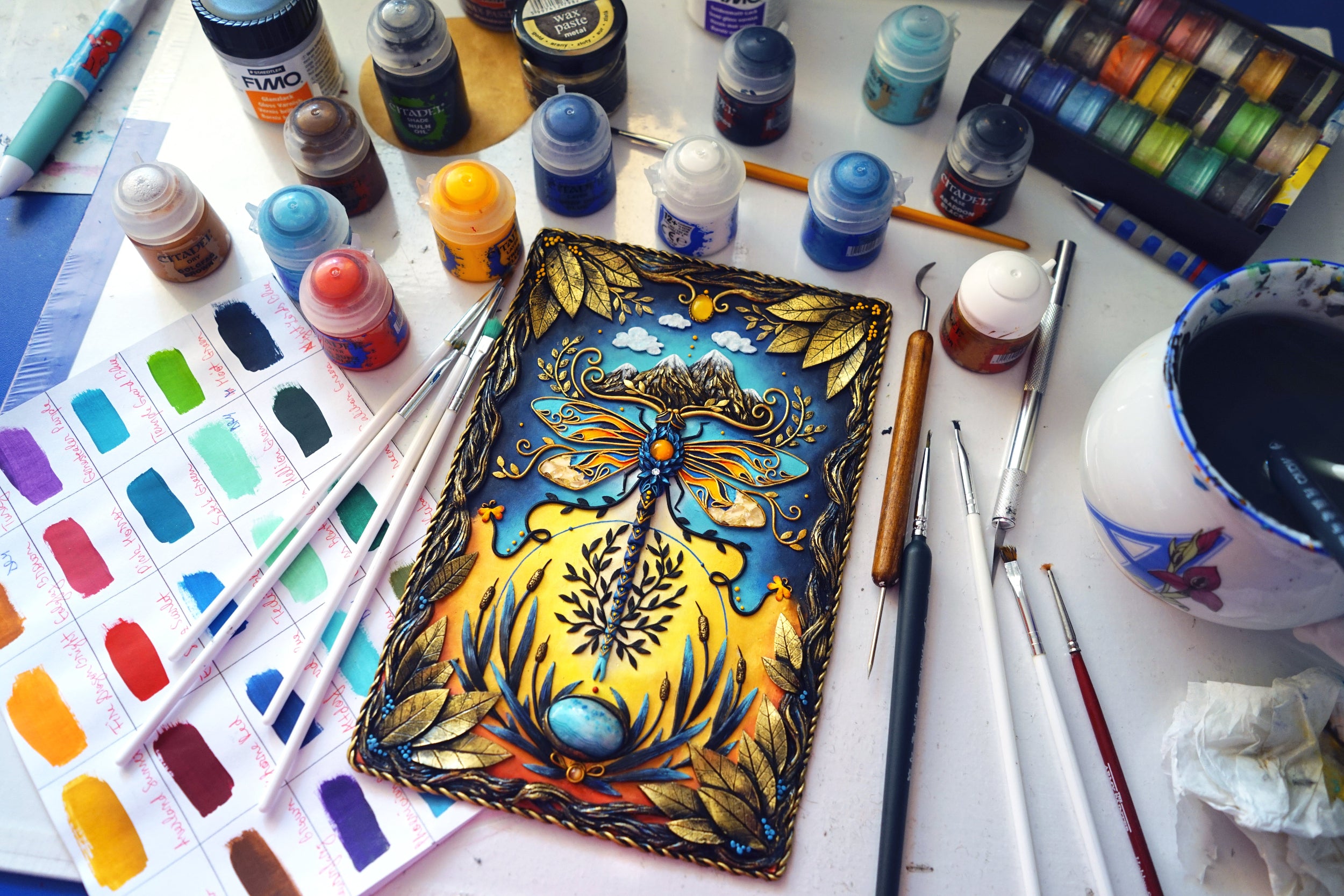 Artistic workspace with painting materials and a colorful dragonfly design made from polymer clay by artist Wildflower.