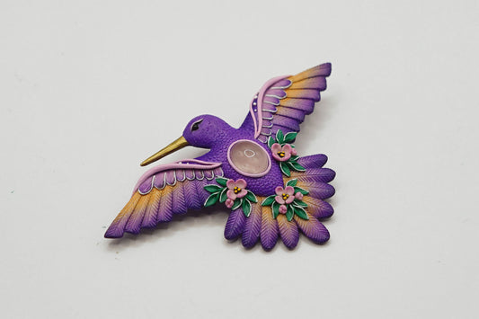 A purple hummingbird-shaped brooch with yellow and orange wing accents, green leaves, pink flowers, and a central circular pearl, shown on a plain light gray background.