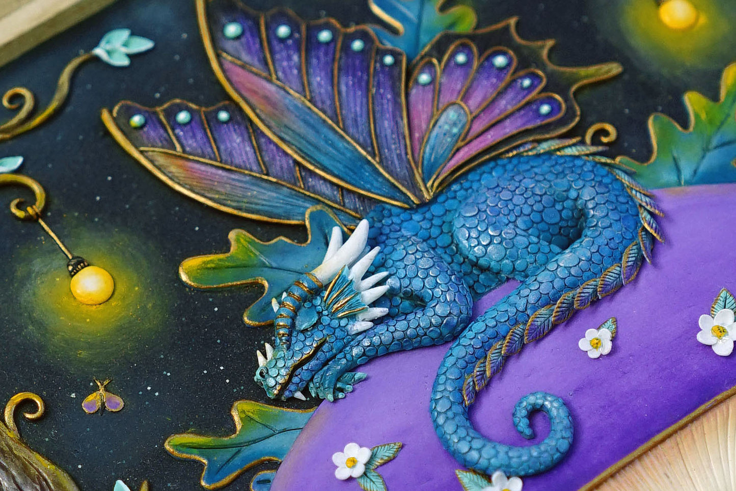 A close-up of a fantasy dragon artwork or decorative panel, showing a blue scaled dragon curled on a purple surface with large purple and teal wings, gold outlines, small white flowers, and glowing yellow lights against a dark starry background.